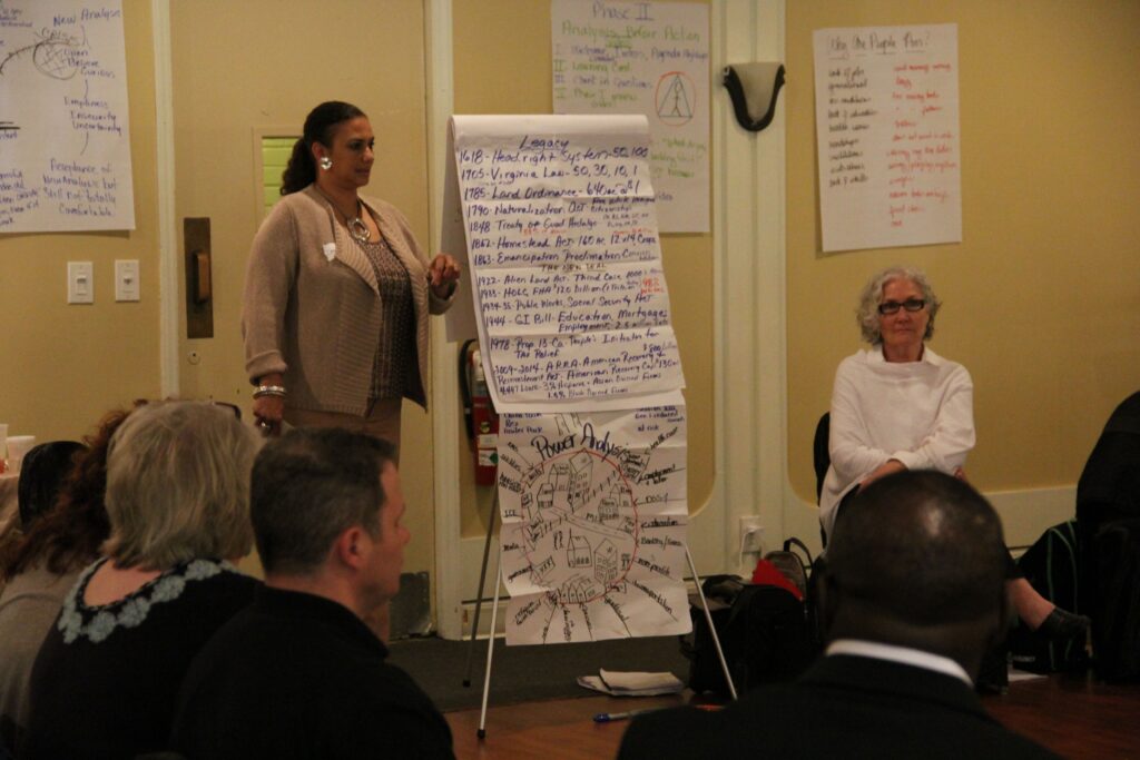 An REI workshop shows participants sitting while one facilitator speaks about the flipchart pages on display. The flipchart pages reflect the group's discussion about "legacy" and "power analysis".